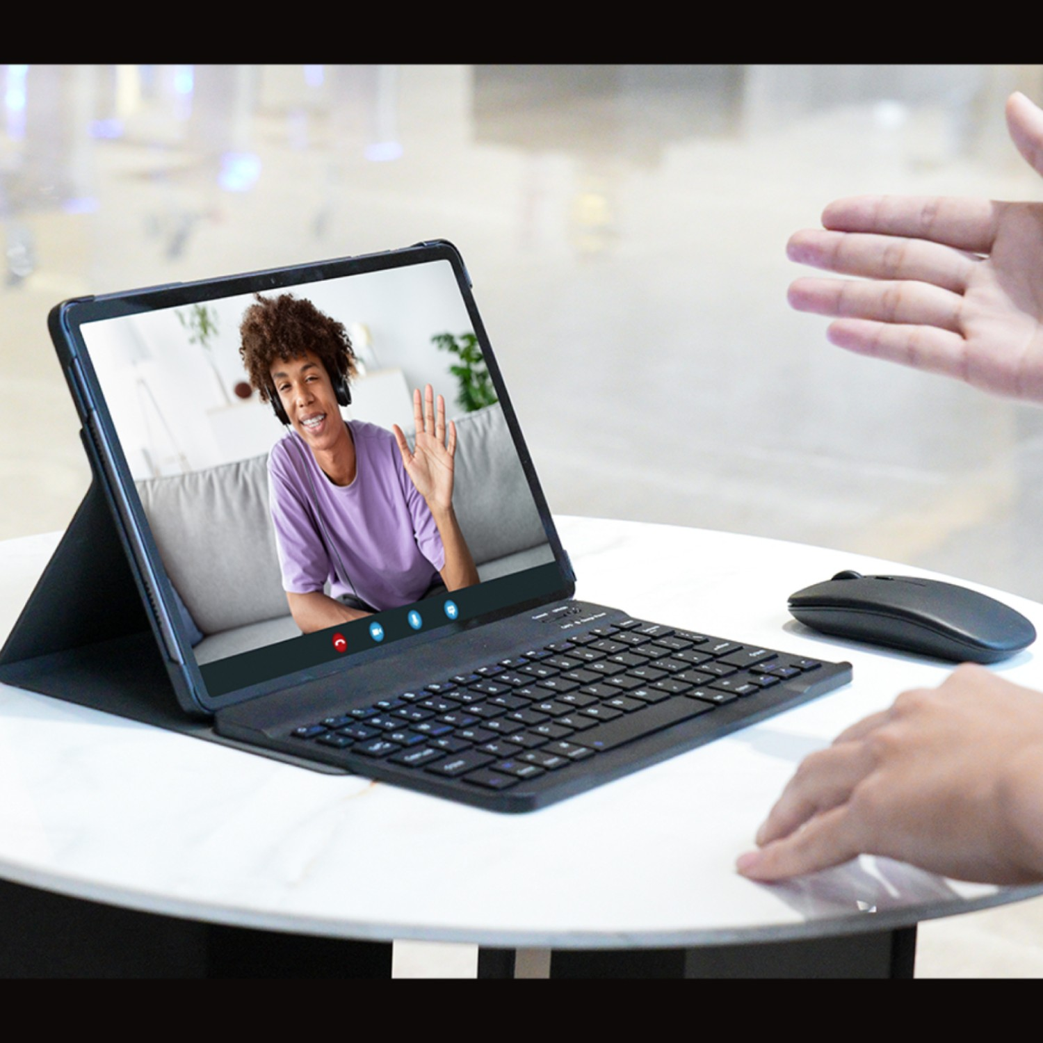 Tablet pokazuje rozmowę wideo. Osoba macha. Klawiatura i mysz na białym stole. Druga ręka sięga.