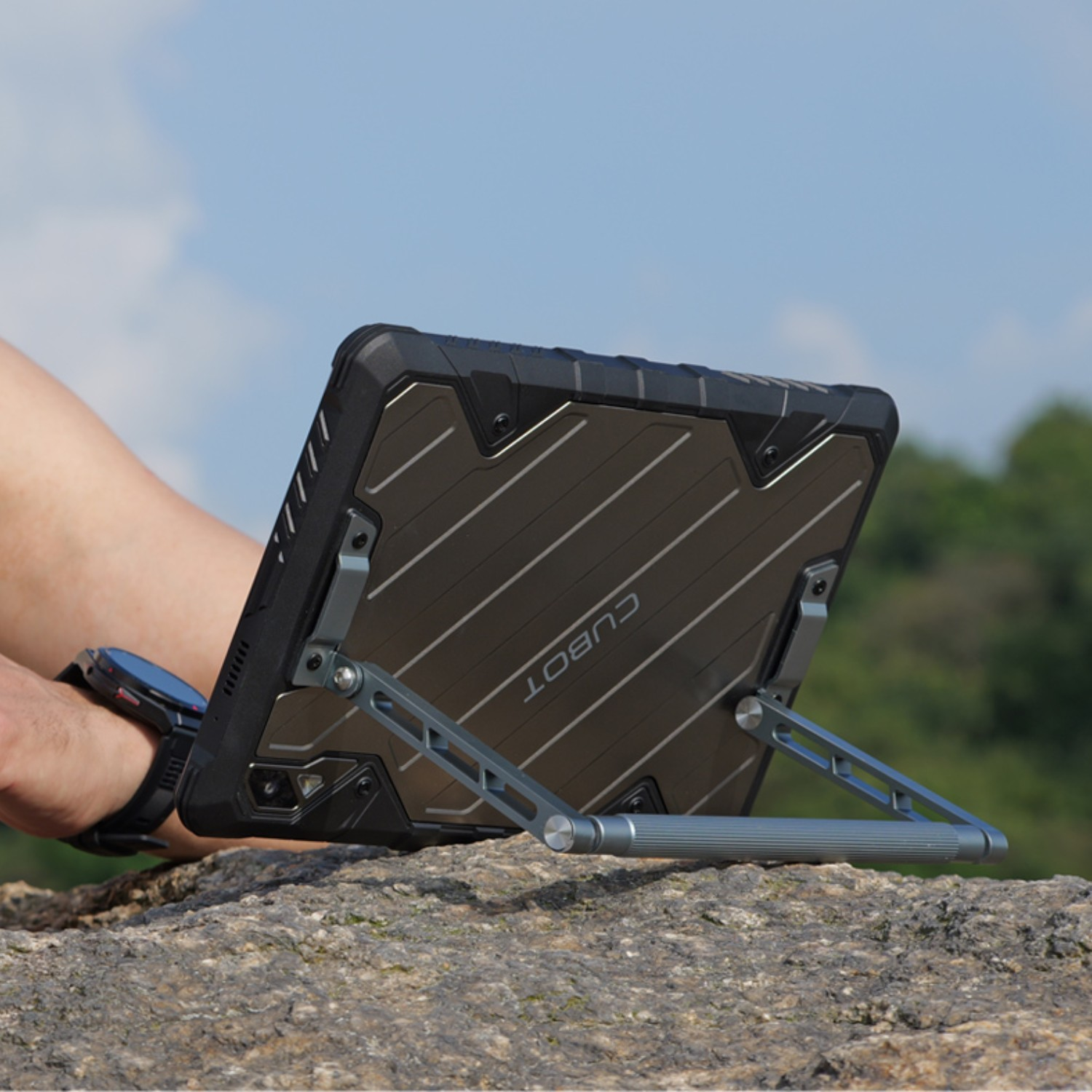 Tablet ze stojakiem na skale. Osoba go trzyma. Błękitne niebo i zielone drzewa w tle. Tablet jest szary.