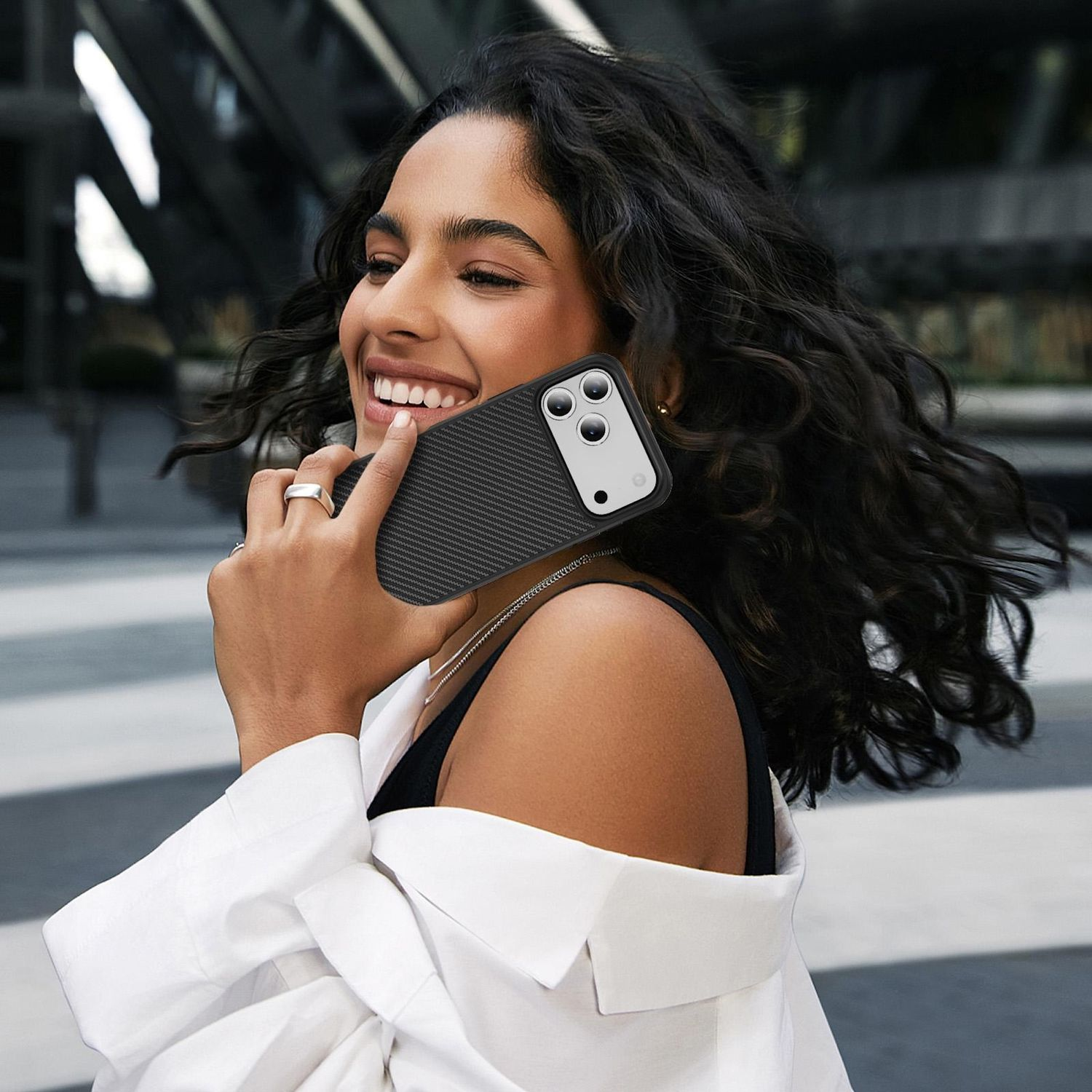 Mujer sonríe, sosteniendo un teléfono en una funda de fibra de carbono. Lleva camisa blanca, anillo plateado y collar. Fondo con edificio.