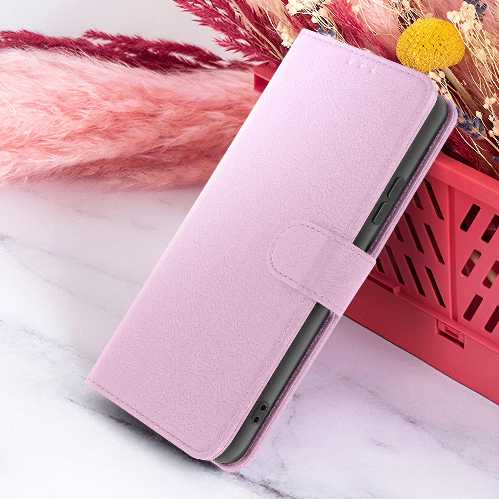Un étui de téléphone rose ouvert, avec un téléphone à l'intérieur. Des fleurs séchées à droite. Une surface rose texturée en arrière-plan.