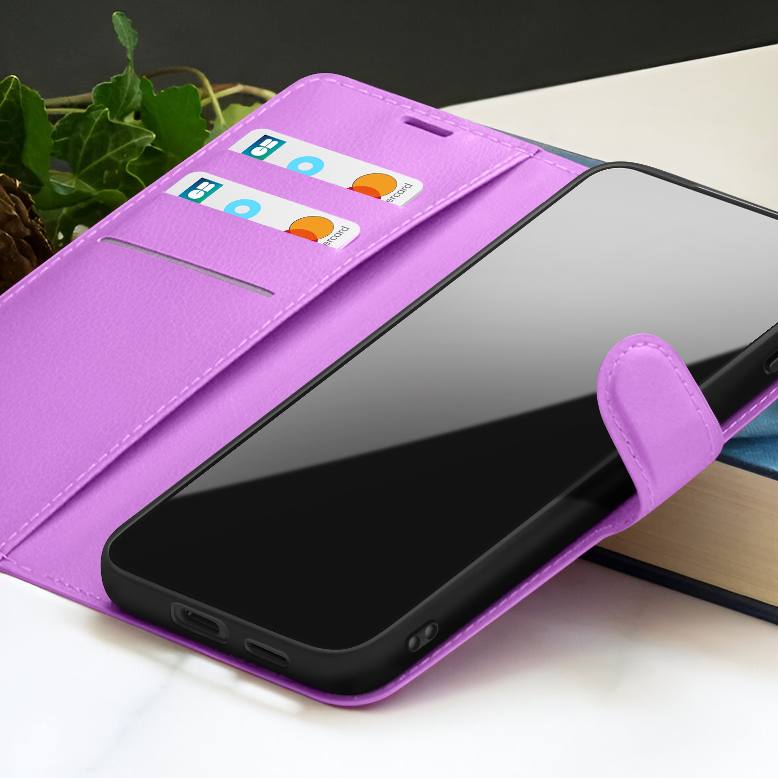 Een smartphone in een paarse leren hoes met creditcardvakjes. Een boek op de achtergrond en groene bladeren links.