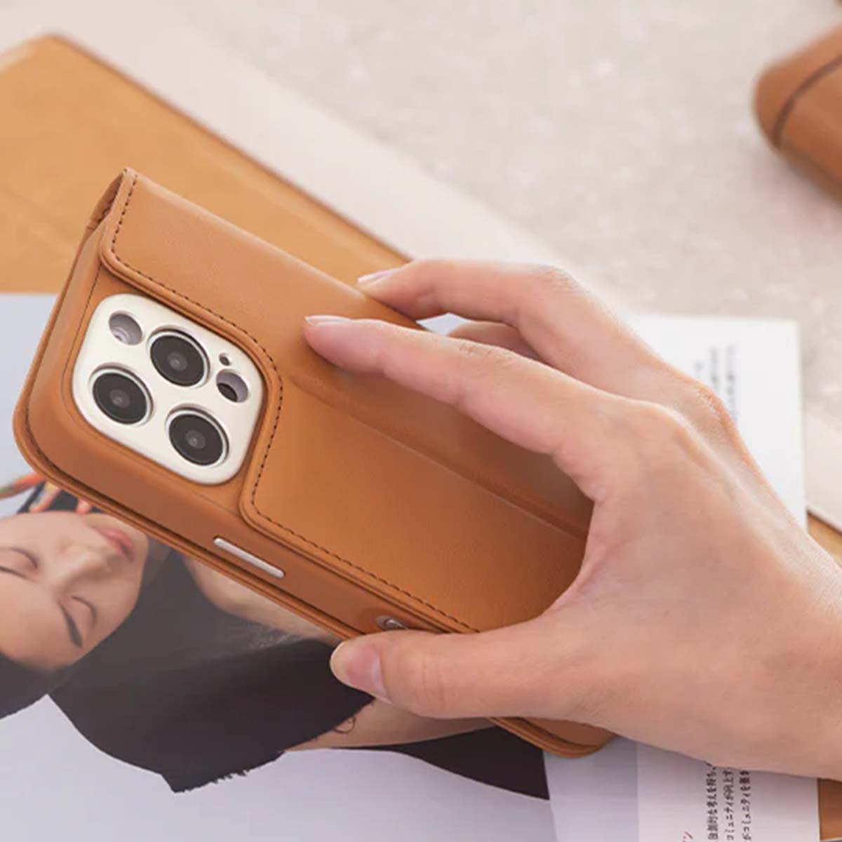 Una mano sostiene una funda de cuero marrón claro sobre una revista. La funda del teléfono está abierta, revelando la cámara del teléfono. El fondo está borroso.