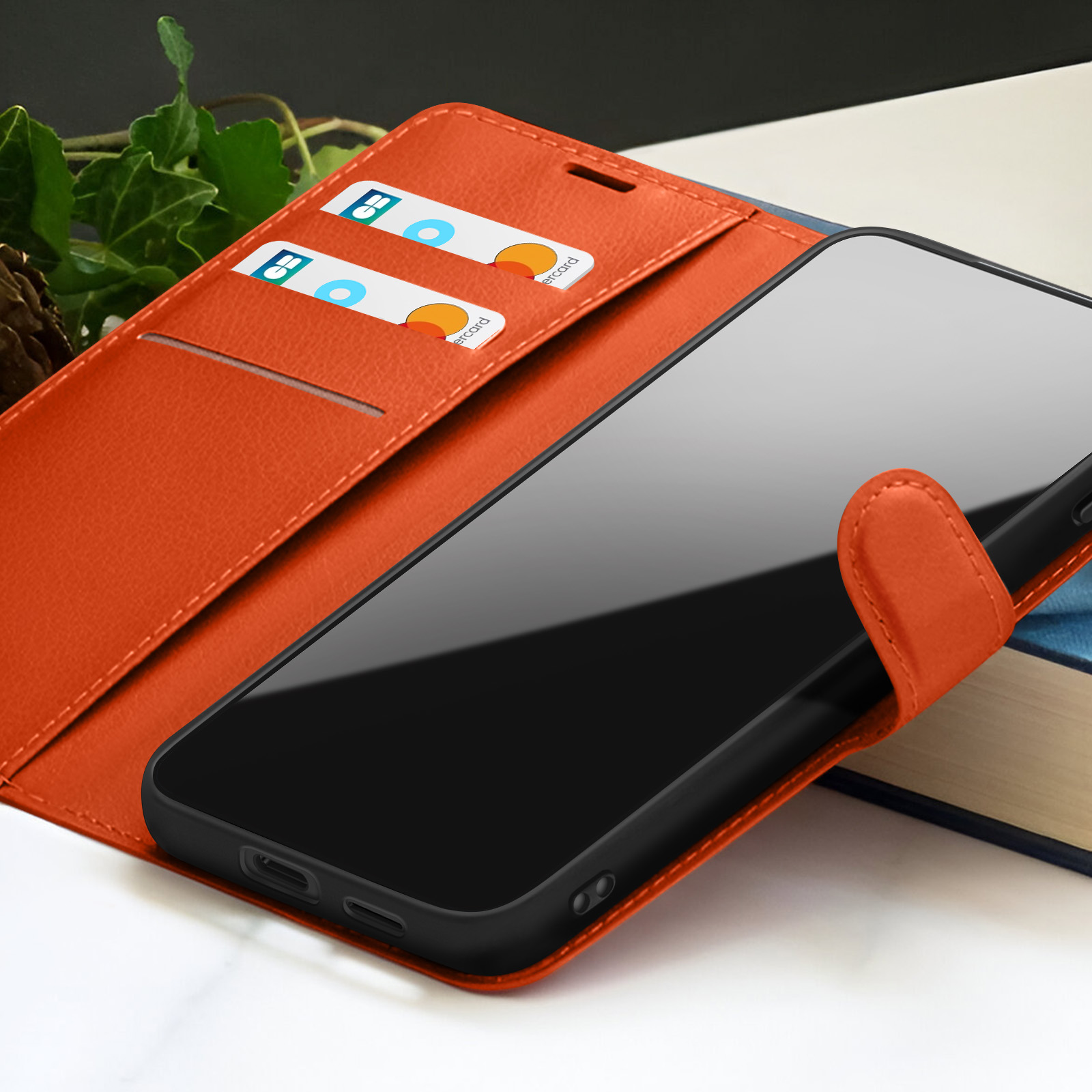 Étui de téléphone orange avec un téléphone noir à l'intérieur. Cartes de crédit dans les fentes. Un livre et des feuilles vertes en arrière-plan.