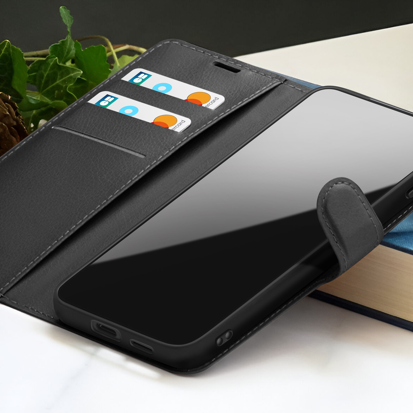 Een zwarte telefoon in een zwarte leren hoes. Creditcards zitten in de hoes. Achtergrond bevat bladeren en een boek.
