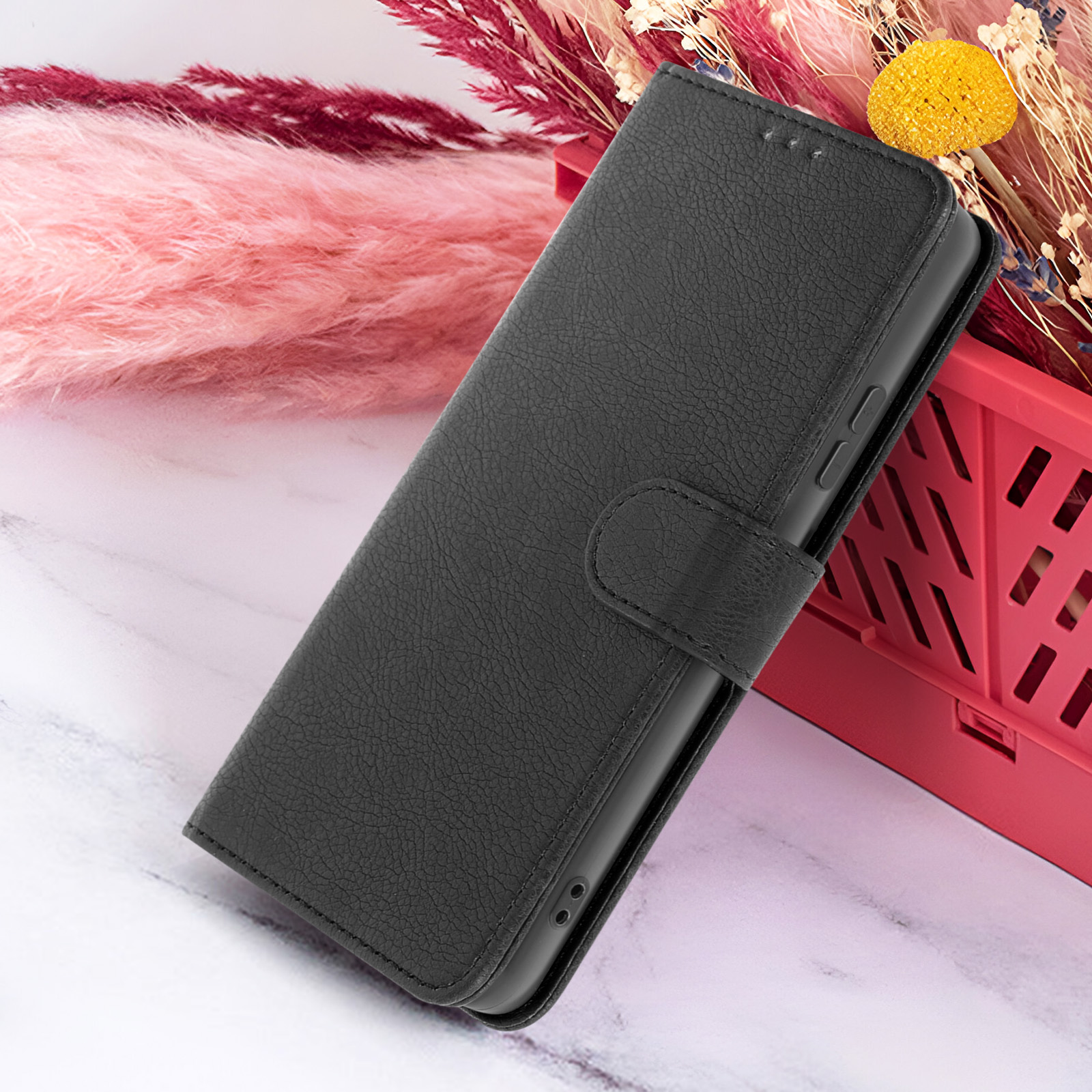 Zwarte leren telefoonhoes met flap, op een wit marmeren oppervlak. Achtergrond met roze en gele decoratieve elementen.