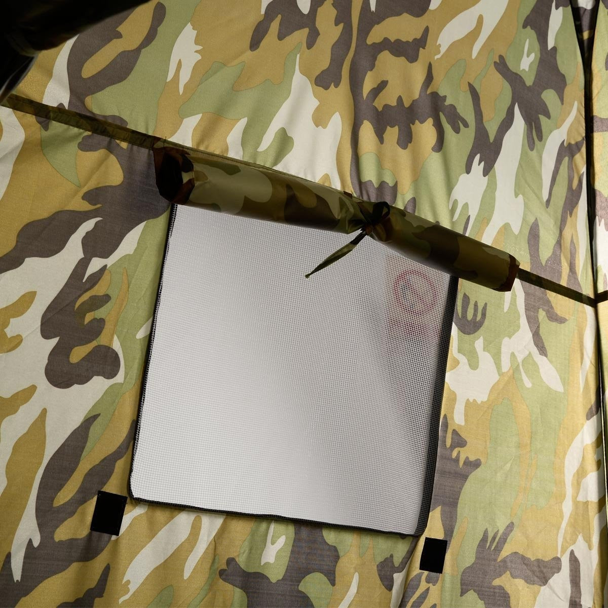 Interior de tienda de camuflaje con ventana de malla. Colores incluyen verde, marrón y beige. Se ven un poste y ataduras.