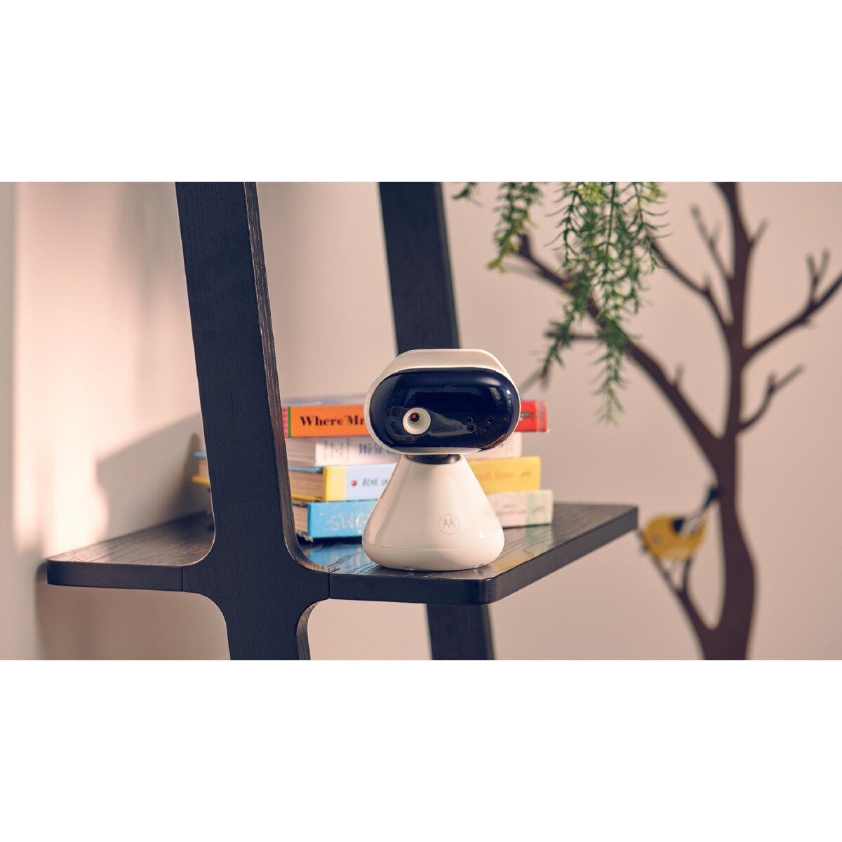 Un baby monitor bianco con lente nera è su uno scaffale con libri. Un albero decorativo e un uccellino sullo sfondo.