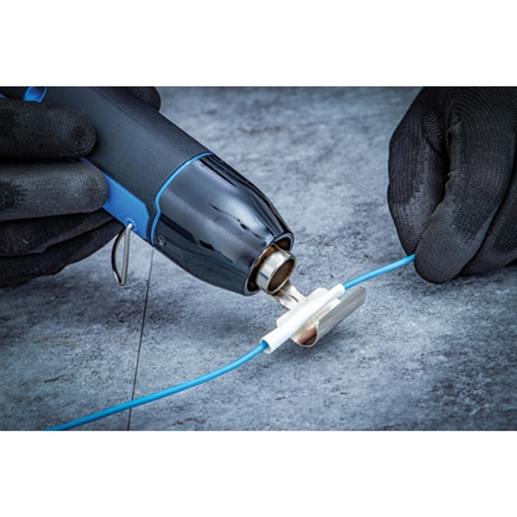 Mani con guanti usano pistola termica su filo blu con connettore bianco, su superficie grigia.