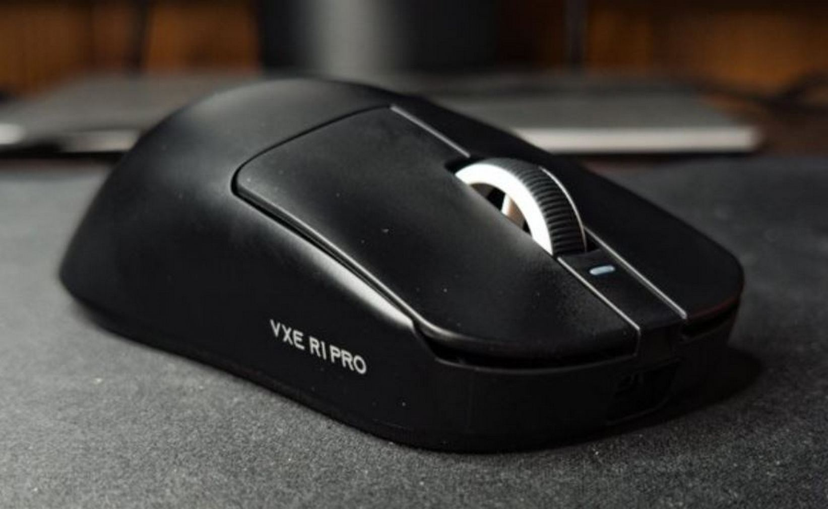 Zwarte VXE R1 Pro draadloze muis op een grijze ondergrond. Witte scrollwiel en blauwe indicator zichtbaar.