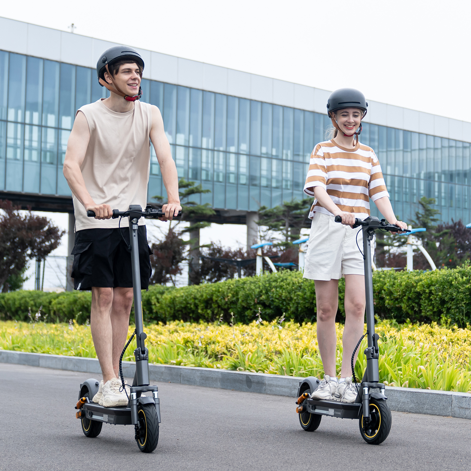 Ein Mann und eine Frau fahren mit Elektrorollern auf einem gepflasterten Weg. Beide tragen Helme. Im Hintergrund ist ein modernes Gebäude zu sehen.