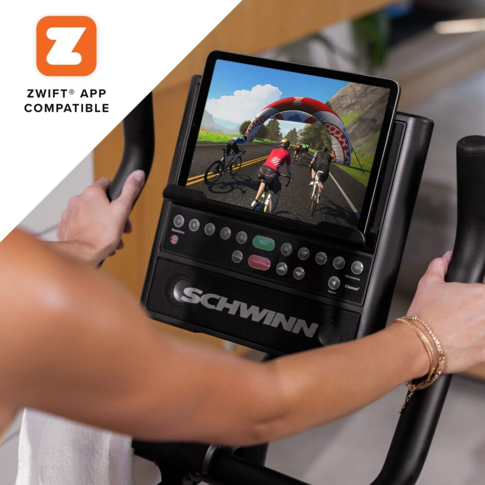 Femme utilisant un vélo d'exercice Schwinn avec une tablette montrant une course cycliste. Mains sur le guidon, bracelets dorés et une serviette blanche.