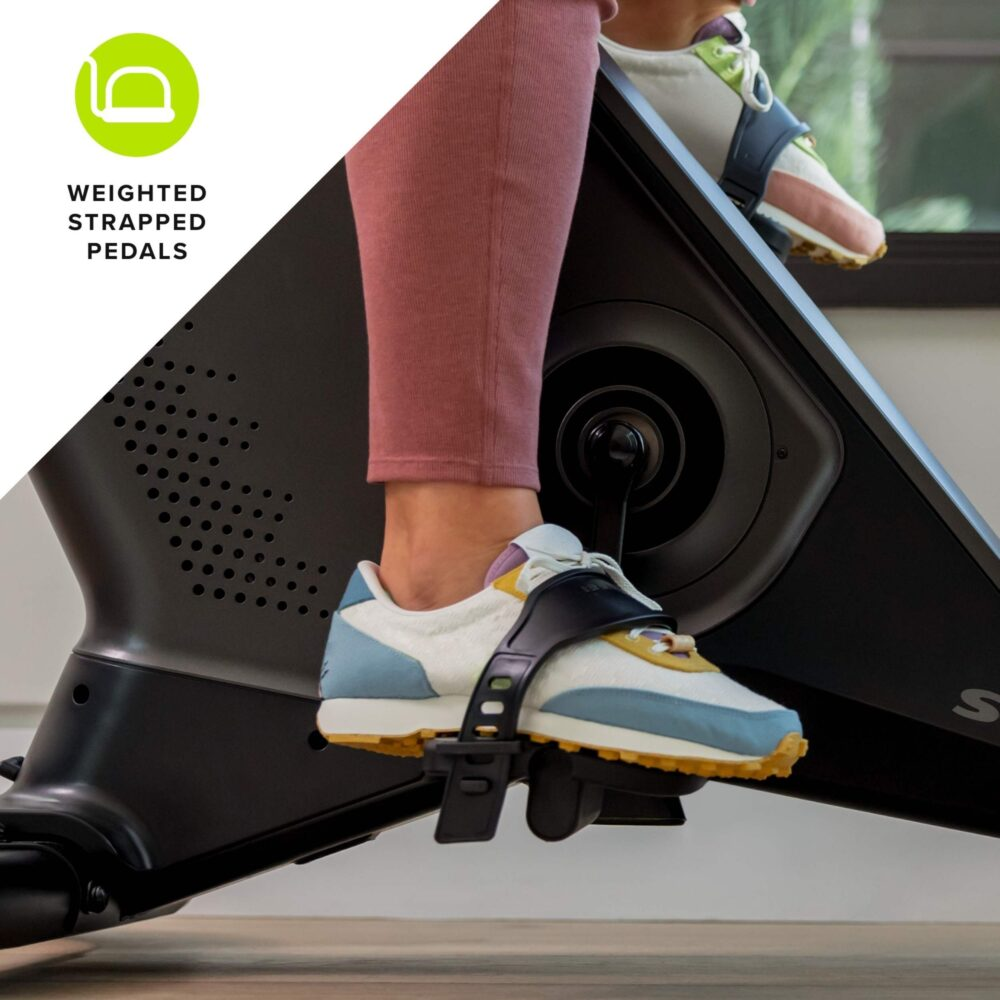 Pieds d'une personne sur un vélo d'appartement, montrant des chaussures attachées aux pédales. Le vélo est noir, avec un legging rose visible.