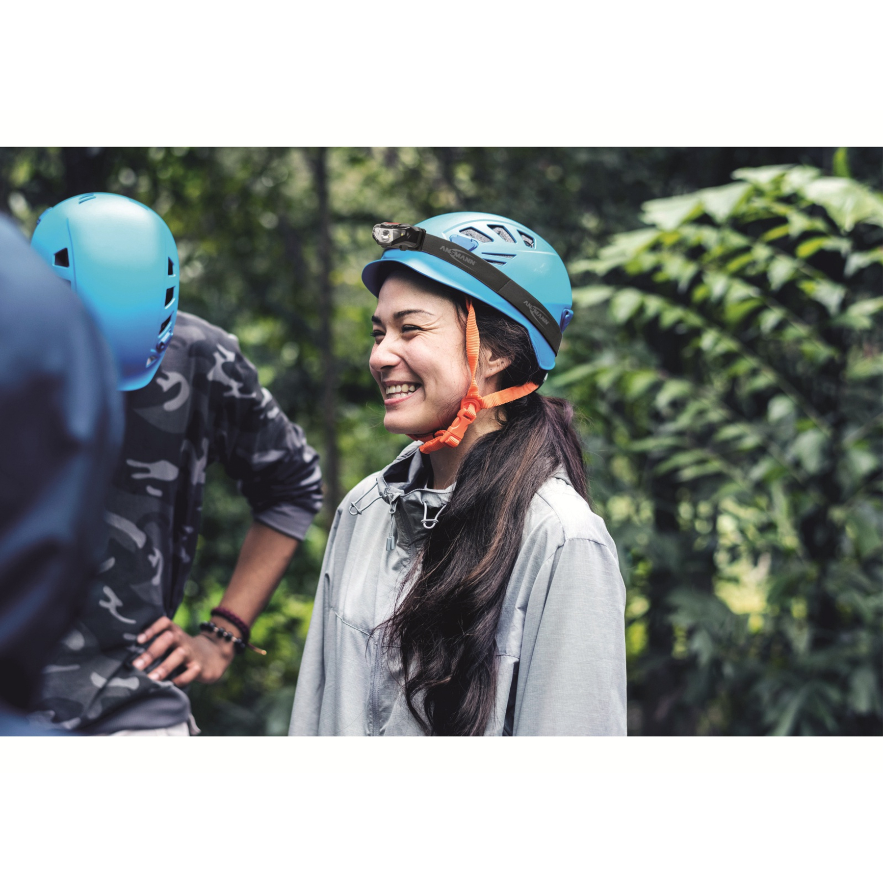 Kobieta z długimi, ciemnymi włosami uśmiecha się, nosząc niebieski kask z czołówką. Druga osoba ma na sobie kask, koszulę moro.