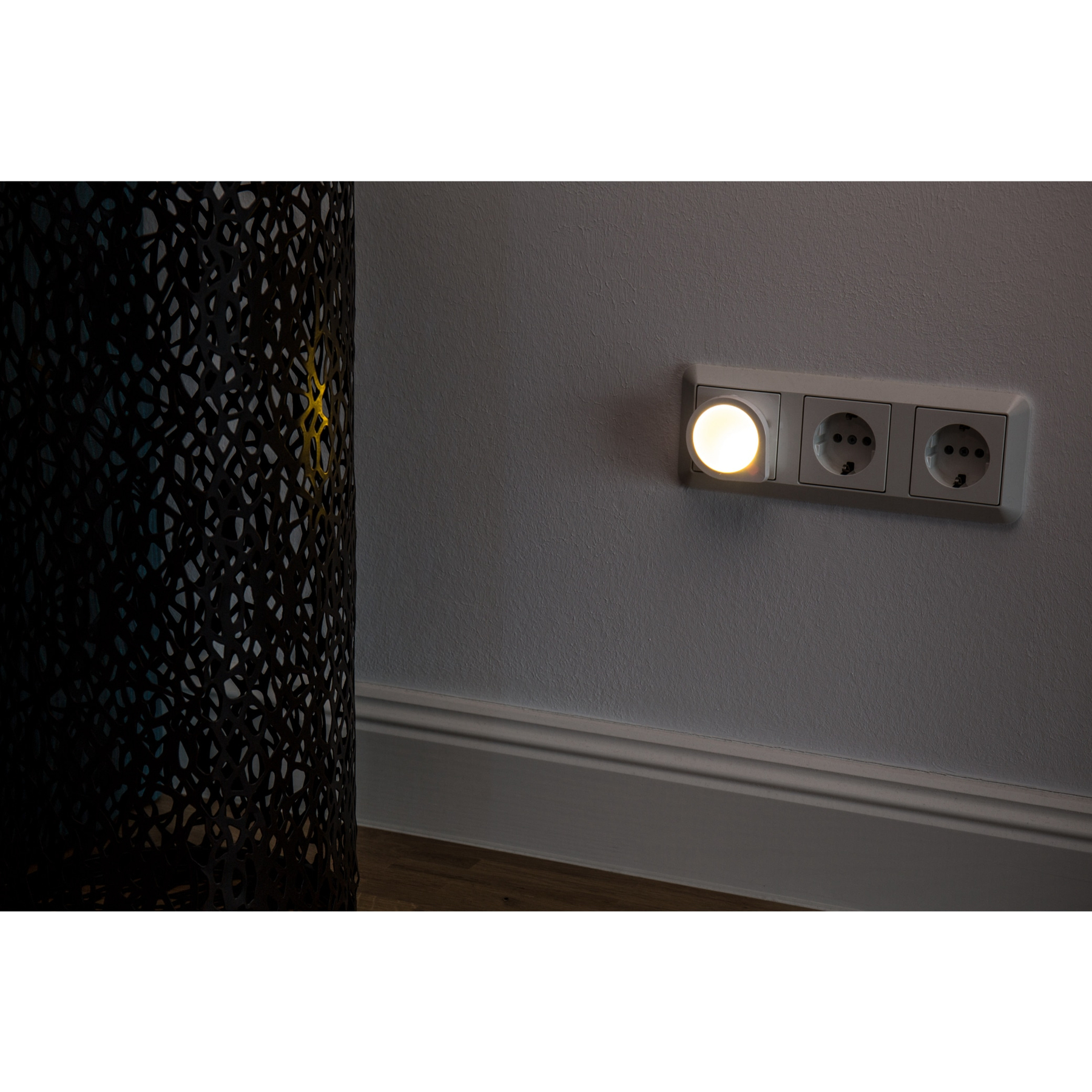 Una presa a muro illuminata con una luce notturna è su un muro grigio. Un oggetto decorativo nero è a sinistra.