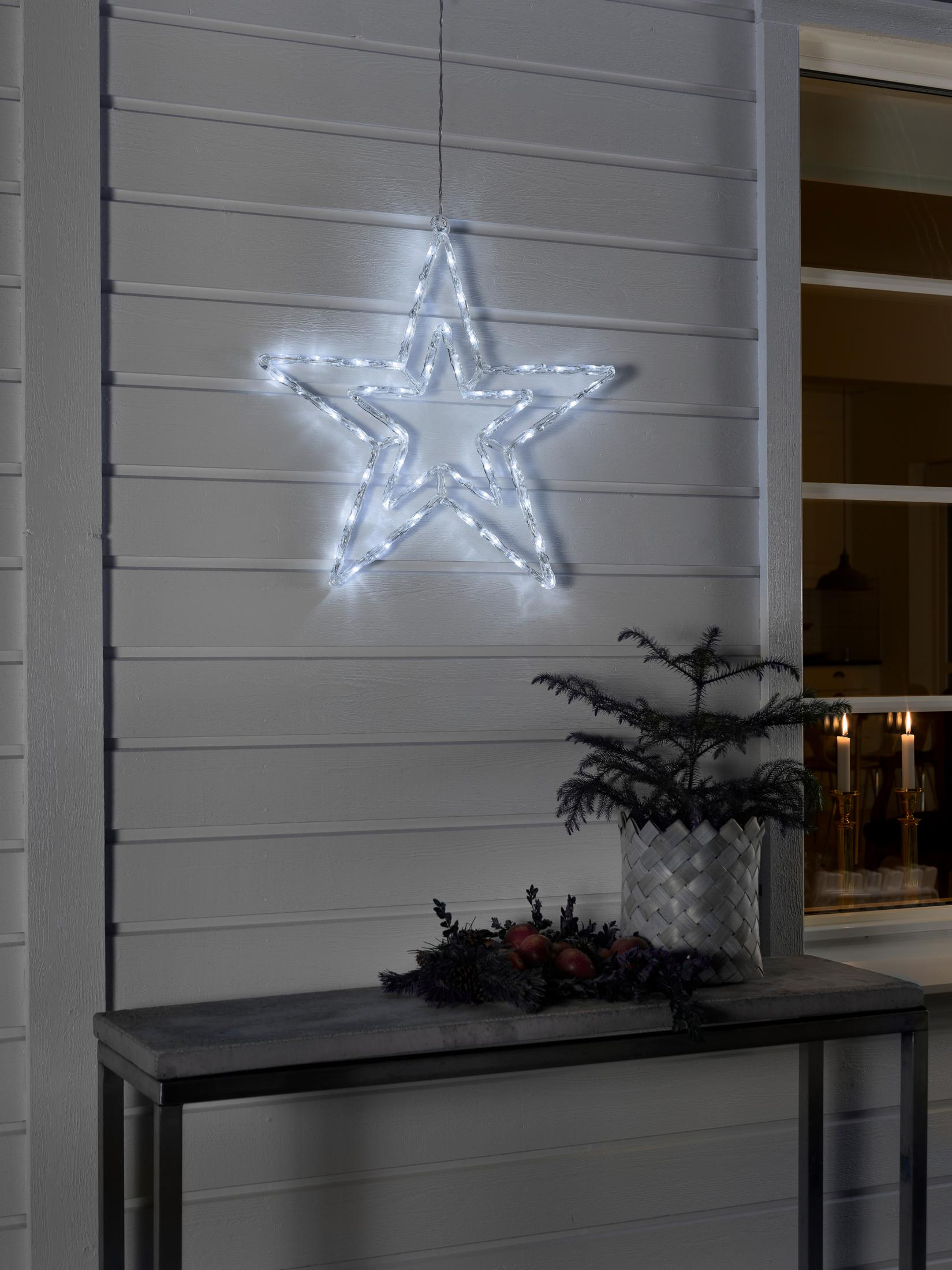 Una decorazione a stella illuminata è appesa a una parete bianca. Un alberello è in un cestino su un tavolo. Ci sono candele nella finestra.