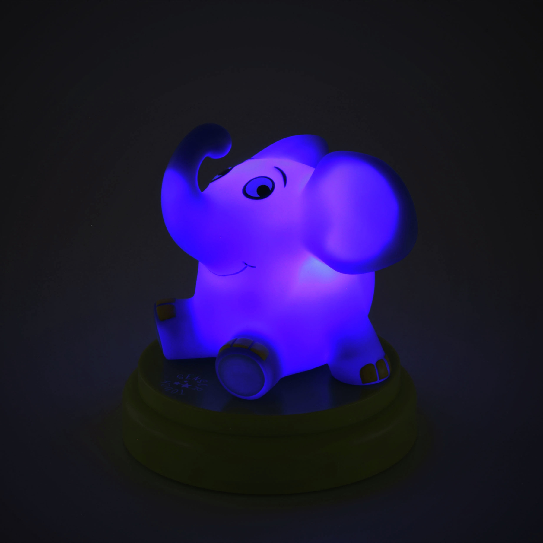 Eine blau leuchtende Elefantenlampe steht auf einem grünen Sockel. Der Hintergrund ist dunkel. Der Elefant hat große Ohren und einen Rüssel.