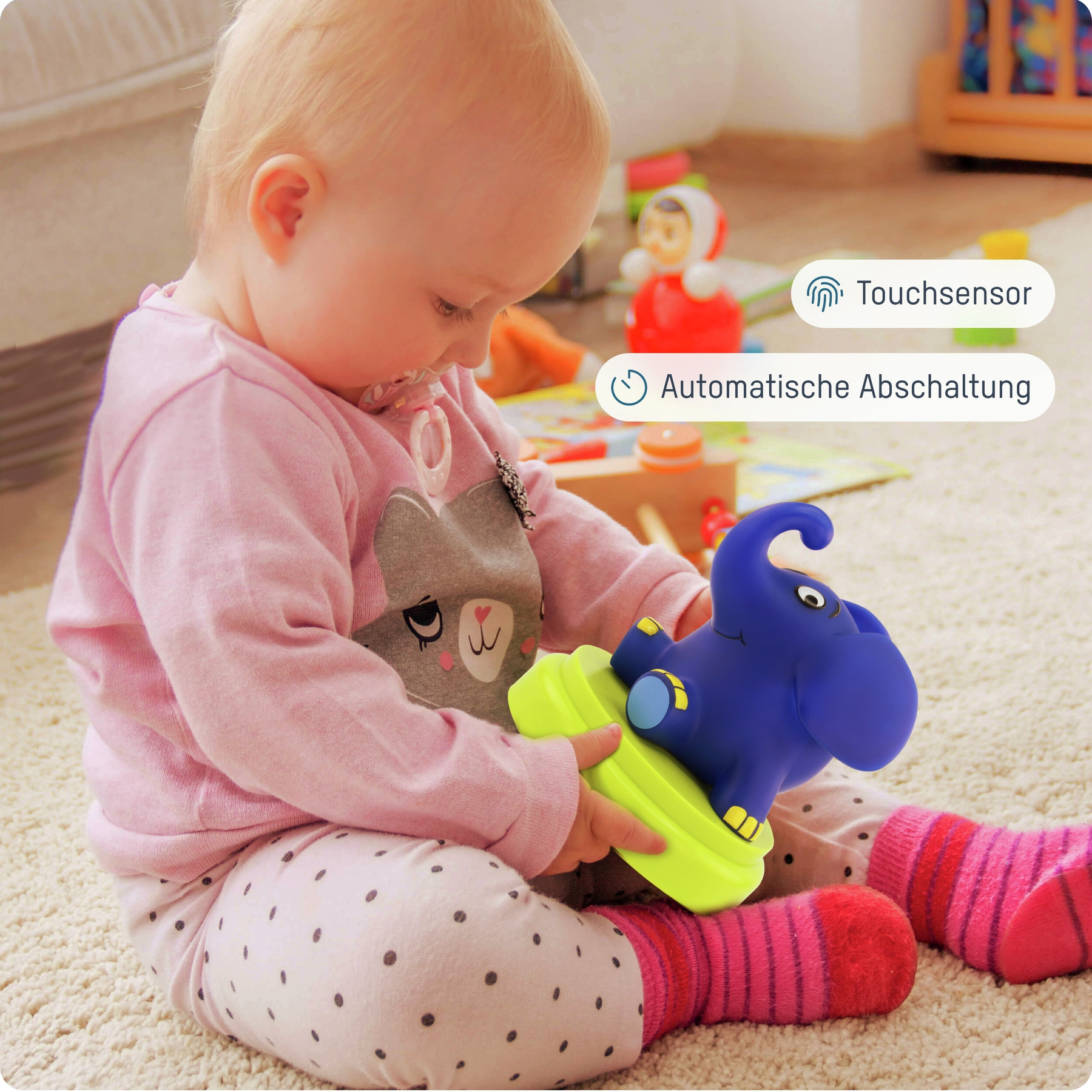 Ein Baby hält ein blaues Elefantenspielzeug mit gelber Basis. Das Baby trägt rosa Kleidung und sitzt auf einem Teppich.