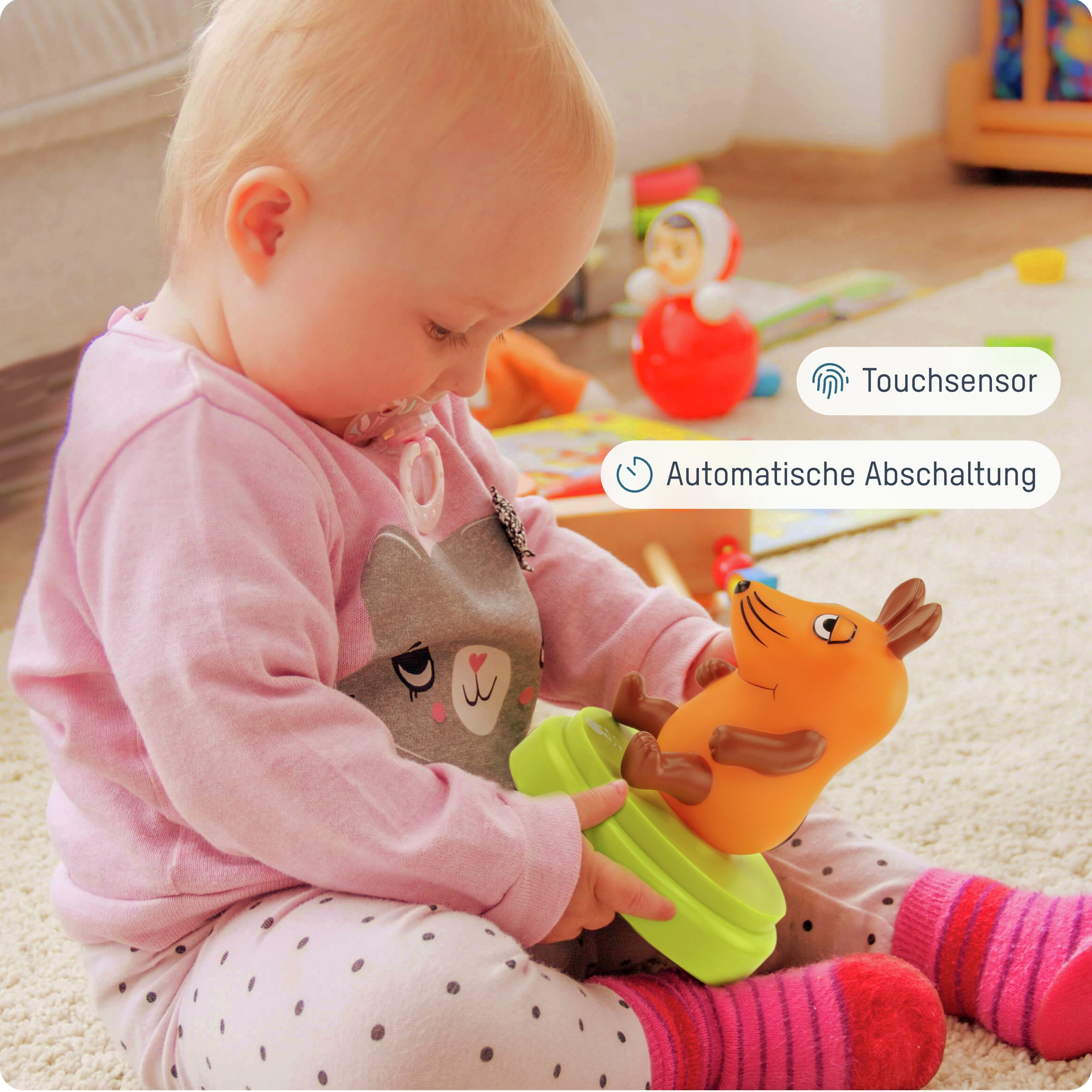 Ein Baby in rosa Kleidung spielt mit einem Spielzeug, das wie eine Cartoon-Maus geformt ist. Andere Spielsachen sind im Hintergrund zu sehen.