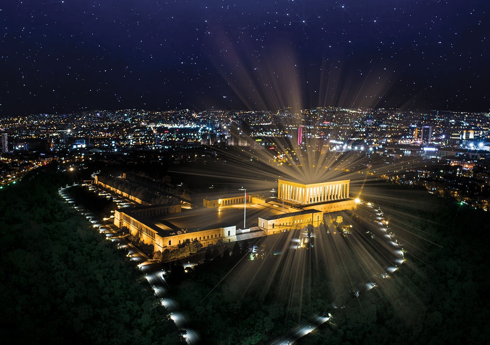 Anıtkabir'in gece havadan görünümü, Ankara, Türkiye, ışık huzmeleriyle aydınlatılmış. Arka planda şehir ı şıkları ve yıldızlı gökyüzü.