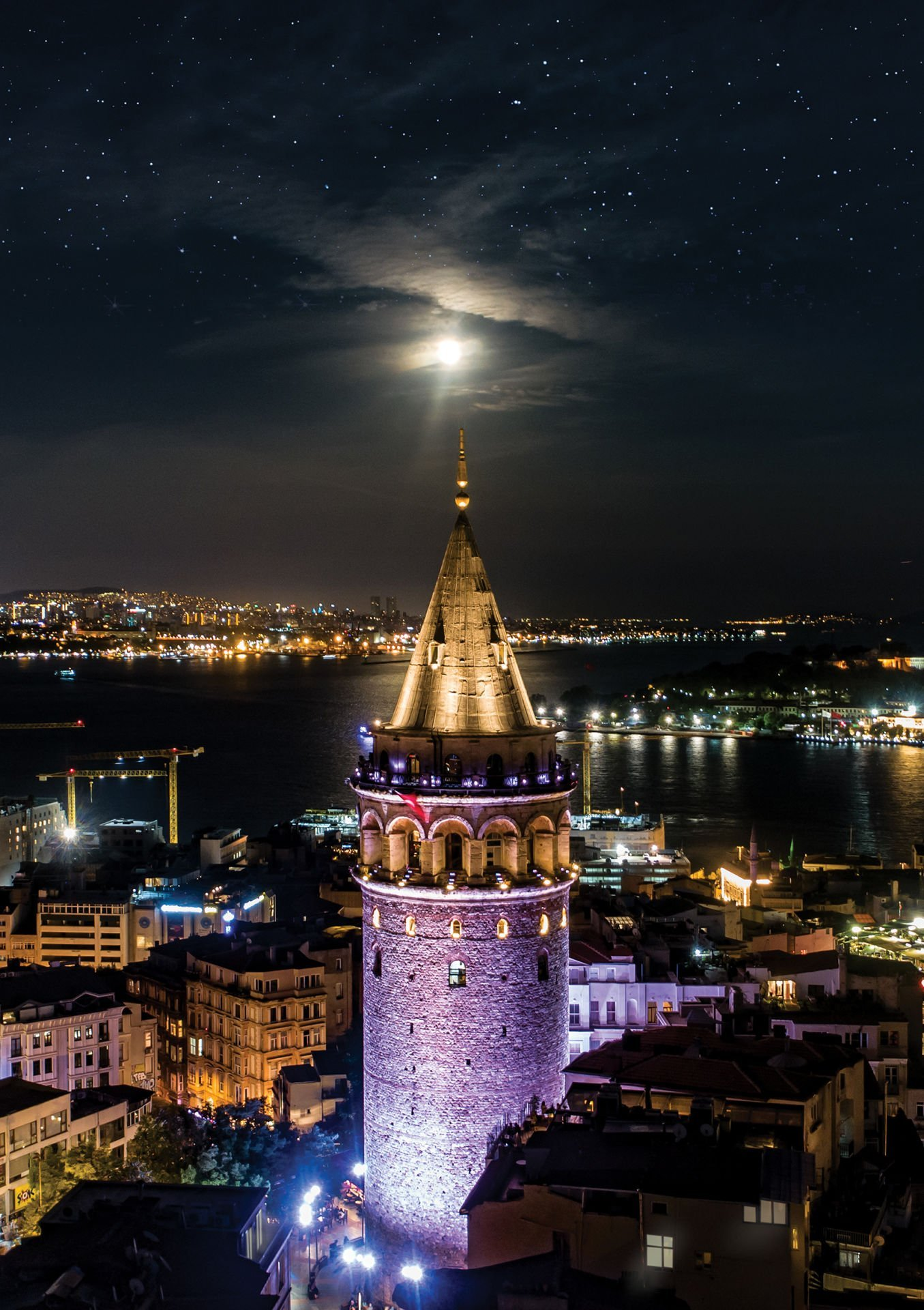 İstanbul'daki Galata Kulesi'nin gece görünümü, Türkiye. Aydınlatılmış kule ve şehir ışıkları suda yansıyor. Ay ve yıldızlar görünür.