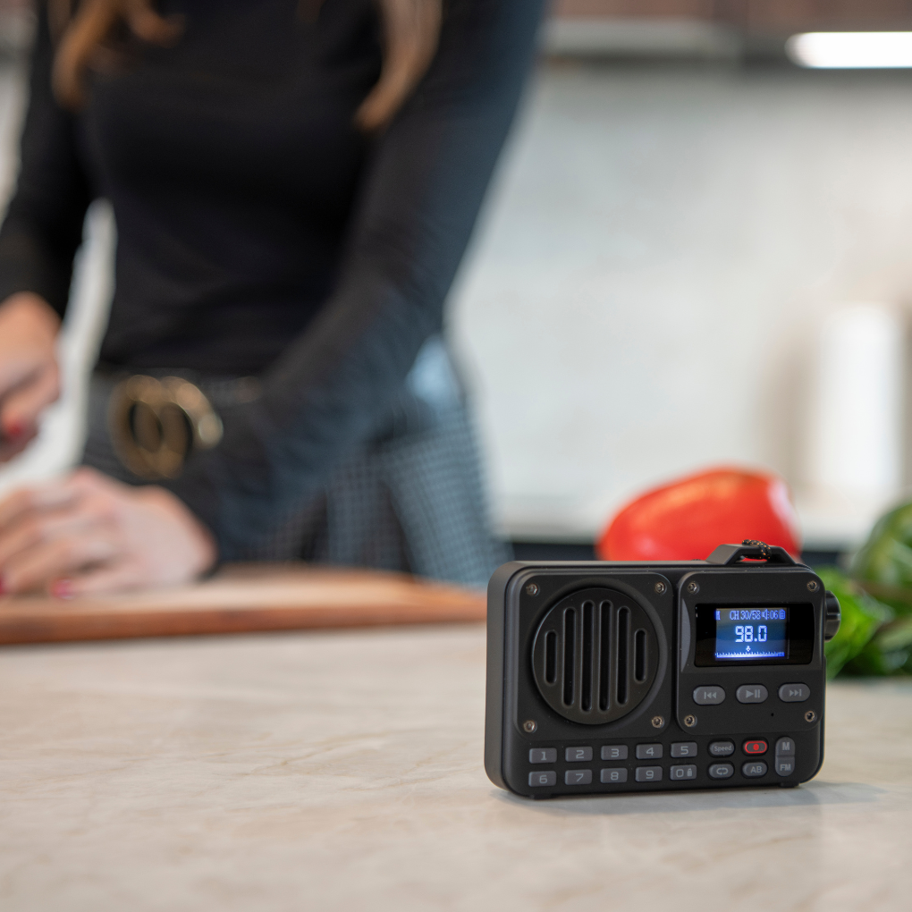 Una radio negra con una pantalla que muestra 98.0 en una mesa. Una persona en el fondo, con un cuchillo cerca de una tabla.