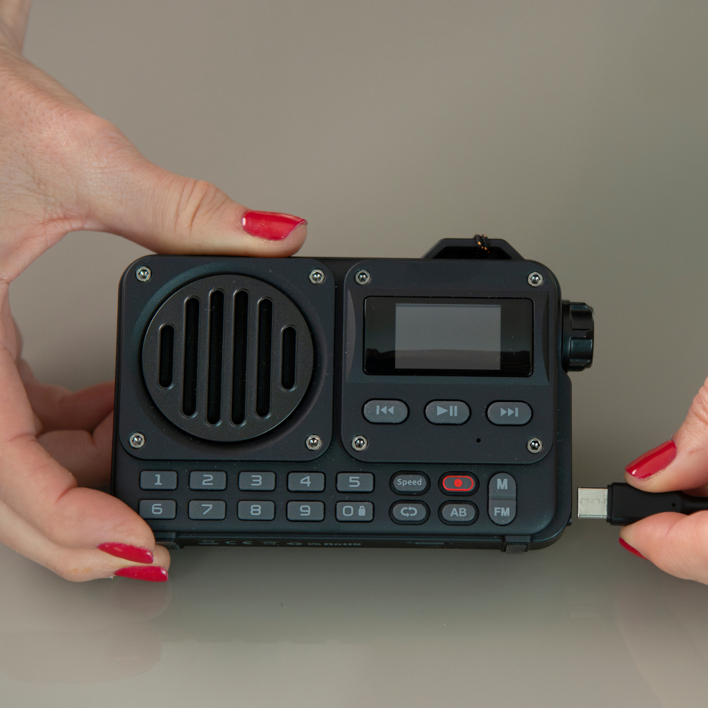 Manos sosteniendo una radio negra, conectando un cable USB. La radio tiene botones y un altavoz. Esmalte de uñas rojo.