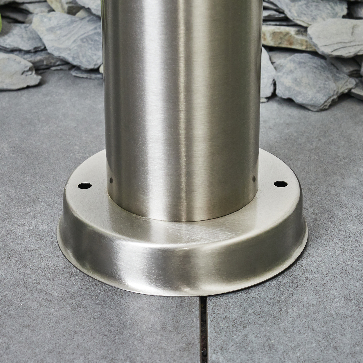 Nahaufnahme einer Stange aus gebürstetem Metall mit kreisförmiger Basis. Die Basis hat zwei kleine Löcher. Der Hintergrund zeigt Steinplatten und eine graue Oberfläche.