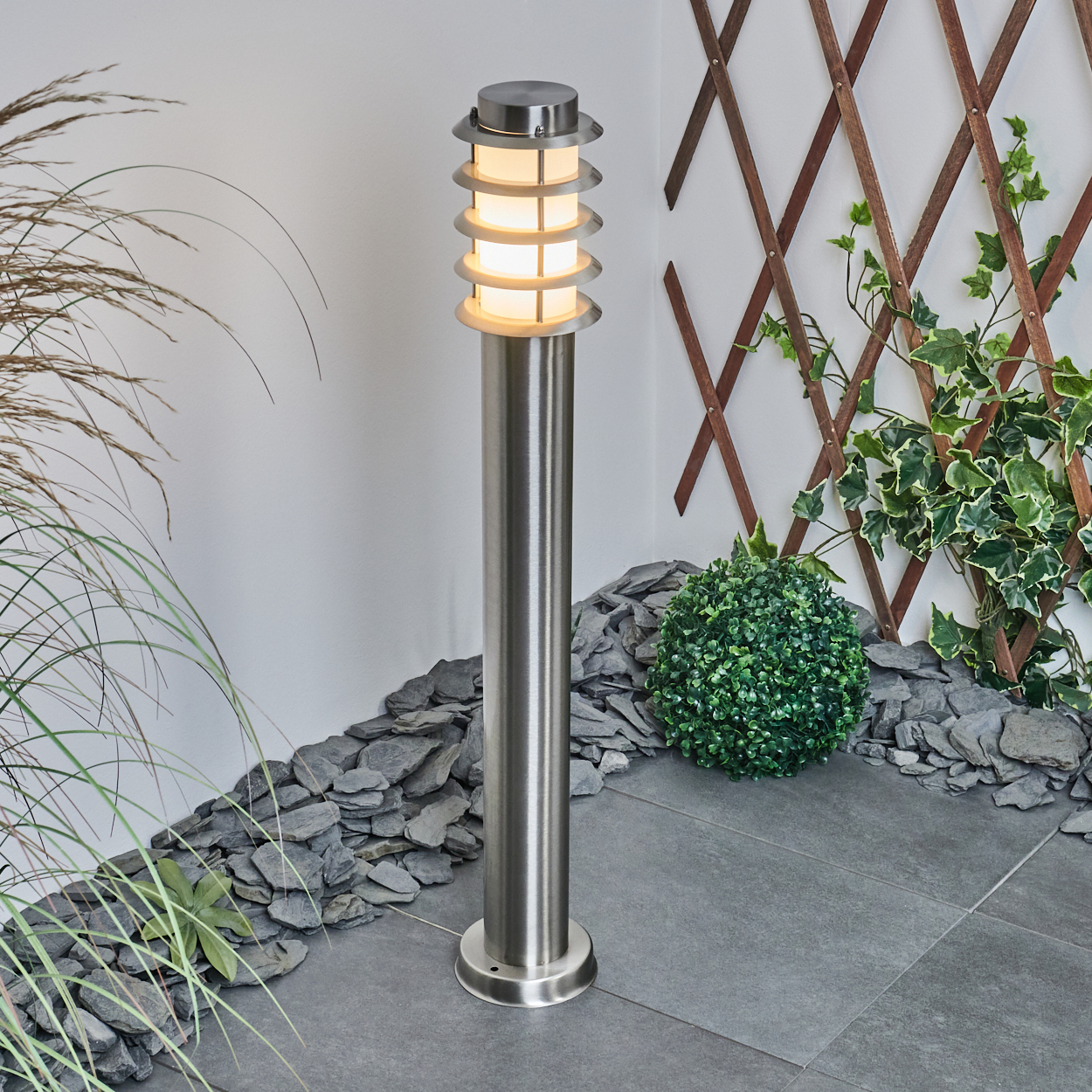 Eine moderne, beleuchtete Außenlampe steht auf einer gefliesten Oberfläche. Die Lampe ist aus Edelstahl, mit Pflanzen und einem Holzgitter im Hintergrund.