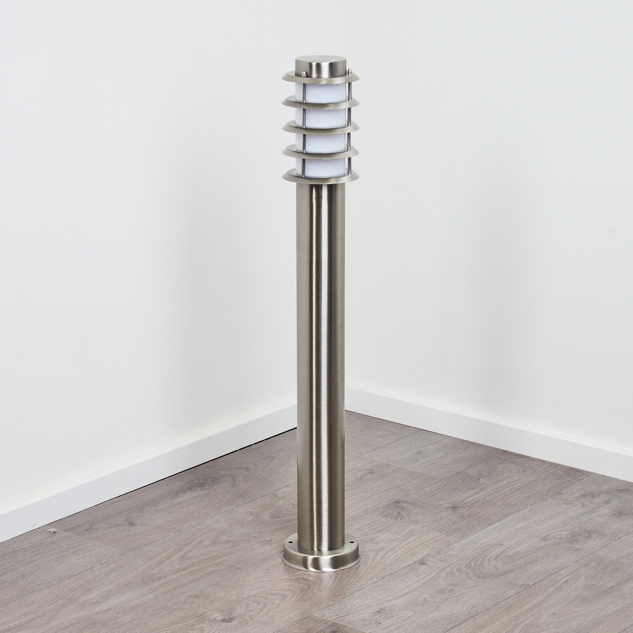 Eine Außenlampe aus Edelstahl steht auf einem Holzboden vor einer weißen Wand. Die Lampe hat eine abgestufte Oberseite und eine zylindrische Basis.