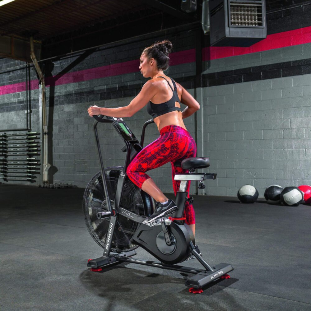 Kobieta ćwiczy na rowerze powietrznym Schwinn w siłowni. Ma na sobie czarny biustonosz sportowy i czerwone legginsy we wzory. Tło ma piłki.