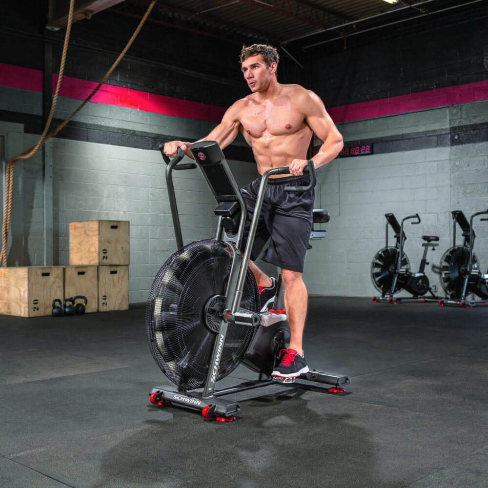 Mężczyzna bez koszuli na rowerze powietrznym, na siłowni. Czarne szorty i buty. Inne rowery treningowe w tle. Drewniane skrzynie i liny.