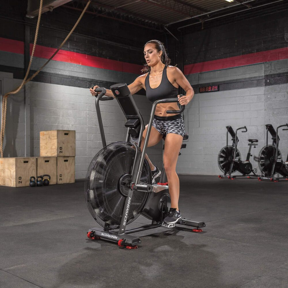 Kobieta ćwiczy na rowerze powietrznym Schwinn na siłowni. Ma na sobie czarny biustonosz sportowy i szorty. Widoczne są inne rowery i drewniane skrzynie.