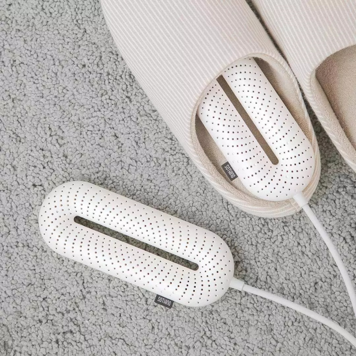 Zwei weiße Schuhtrockner mit Perforationen liegen auf einem grauen Teppich, einer davon in einem hellen Hausschuh.