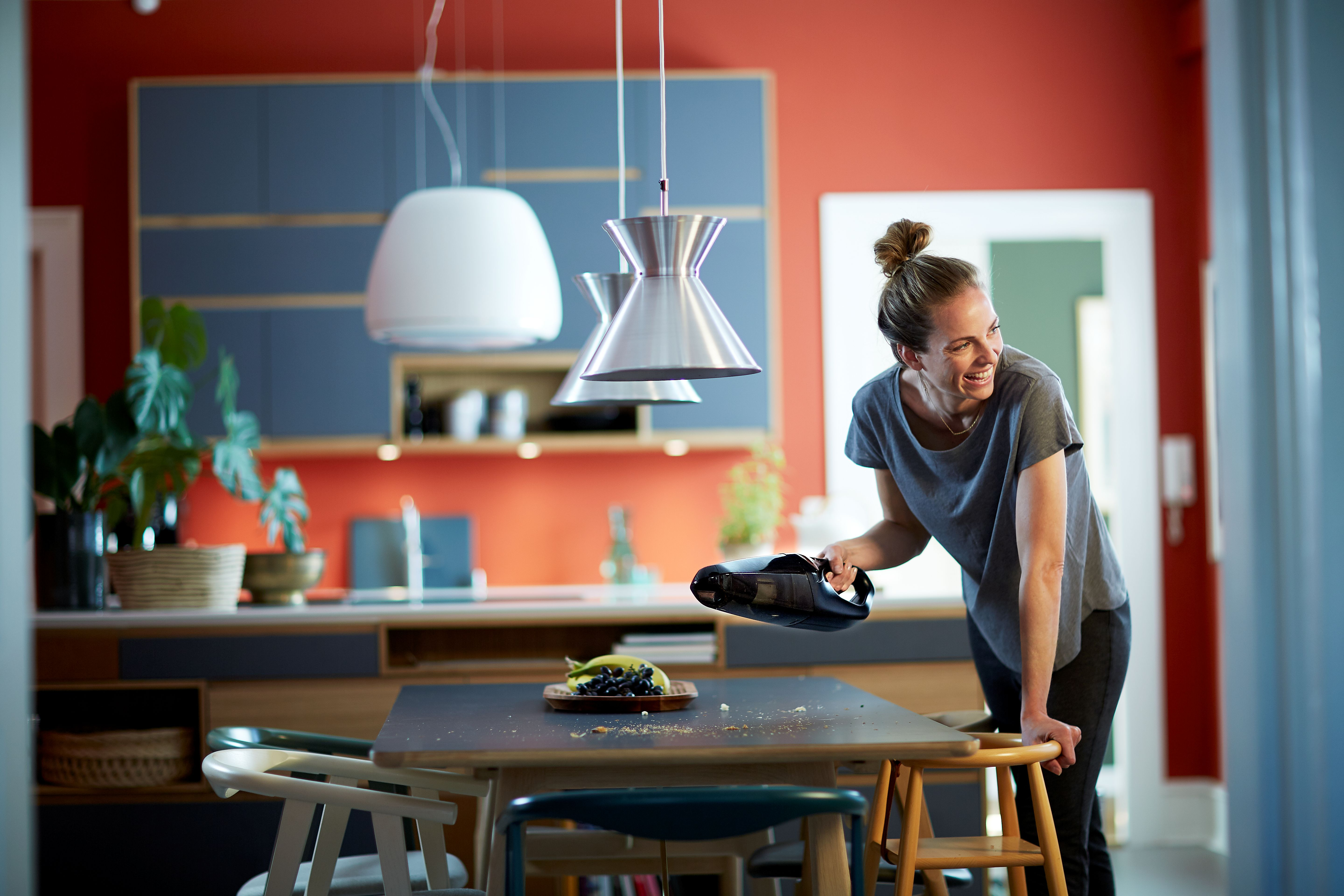 Woman smiling, holding a vacuum cleaner in a modern kitchen. Table with fruit and plants in the background.