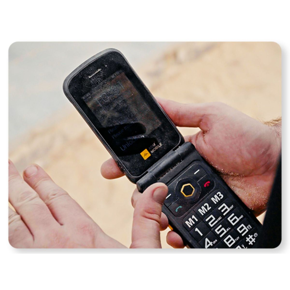 Eine Person hält ein schwarzes Klapphandy. Das Telefon ist geöffnet und zeigt einen Bildschirm. Der Hintergrund ist eine verschwommene, sandige Textur.