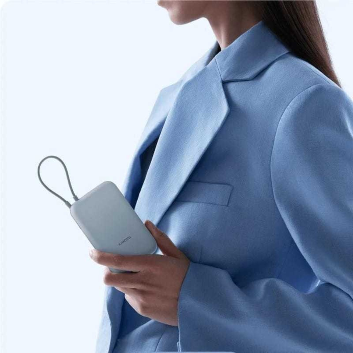 Osoba trzyma jasnoniebieski power bank. Ma na sobie jasnoniebieską marynarkę. Tło jest jednolite, jasnoniebieskie.