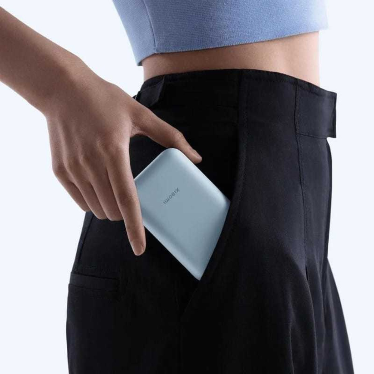 Ręka osoby wkładająca jasnoniebieski power bank do czarnej kieszeni spodni. Osoba nosi jasnoniebieski top.