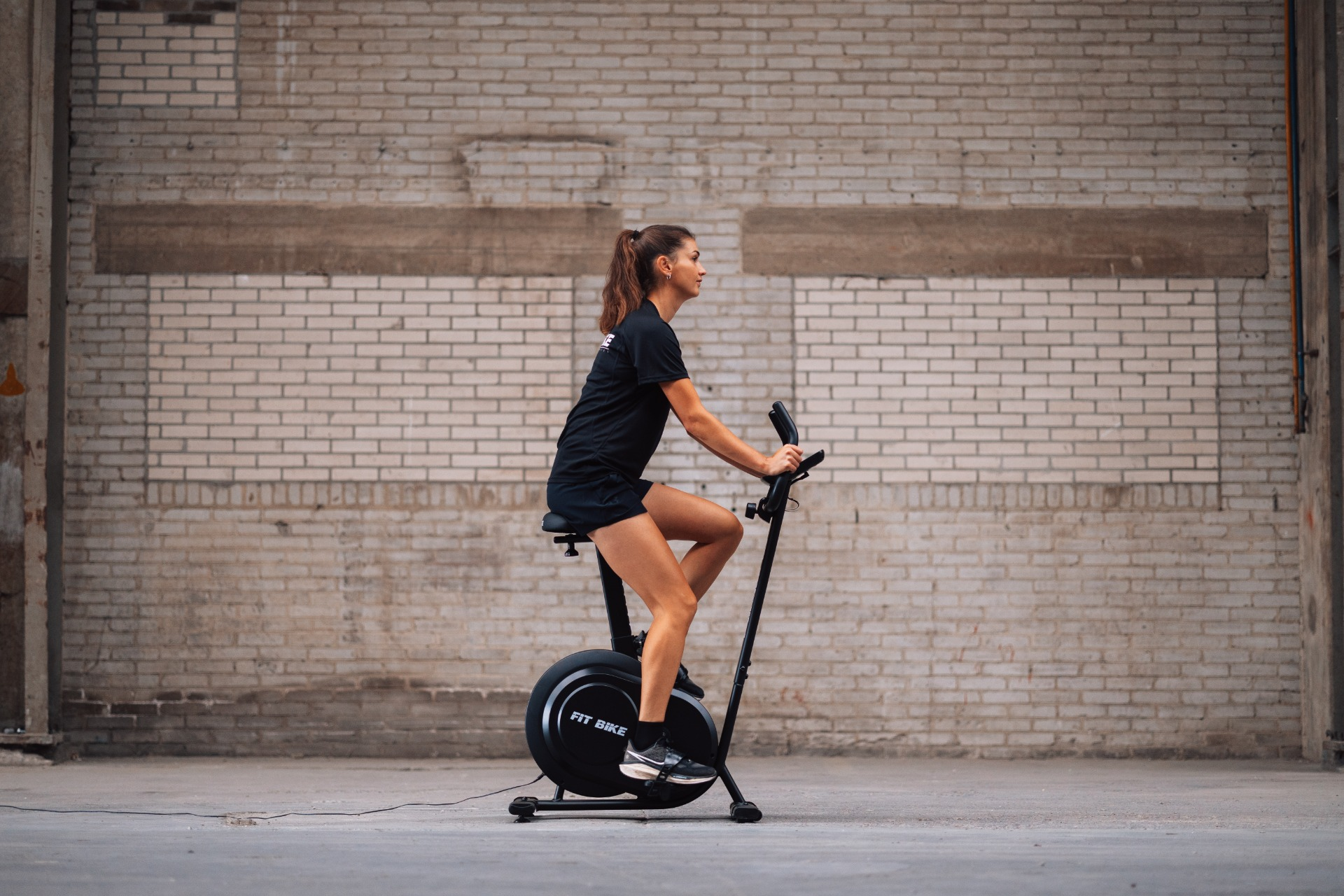 Woman on a stationary bike in front of a brick wall. She wears black athletic wear.