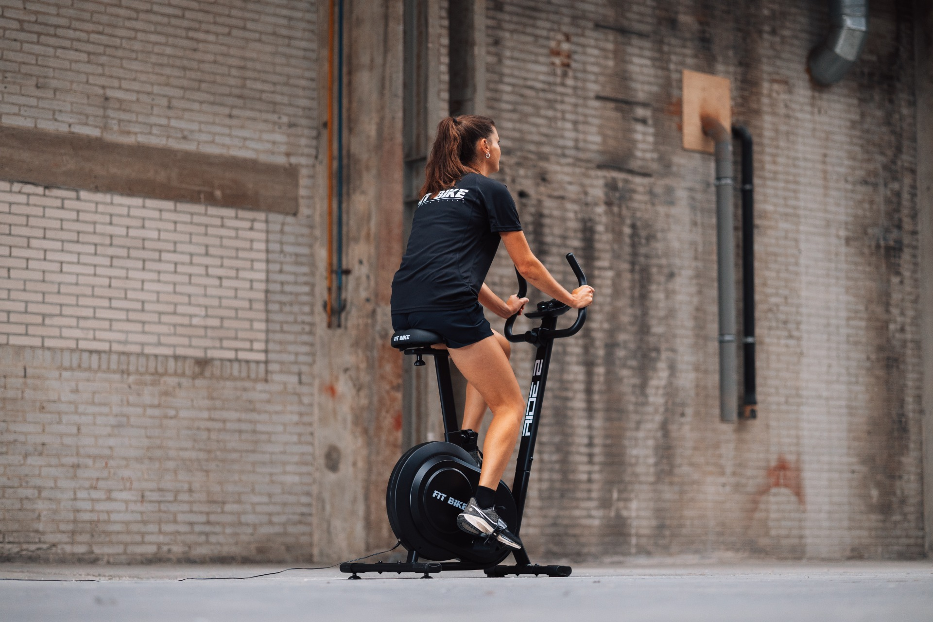 Woman on a stationary bike in front of a brick wall. She is wearing a black shirt and shorts. The bike is black.