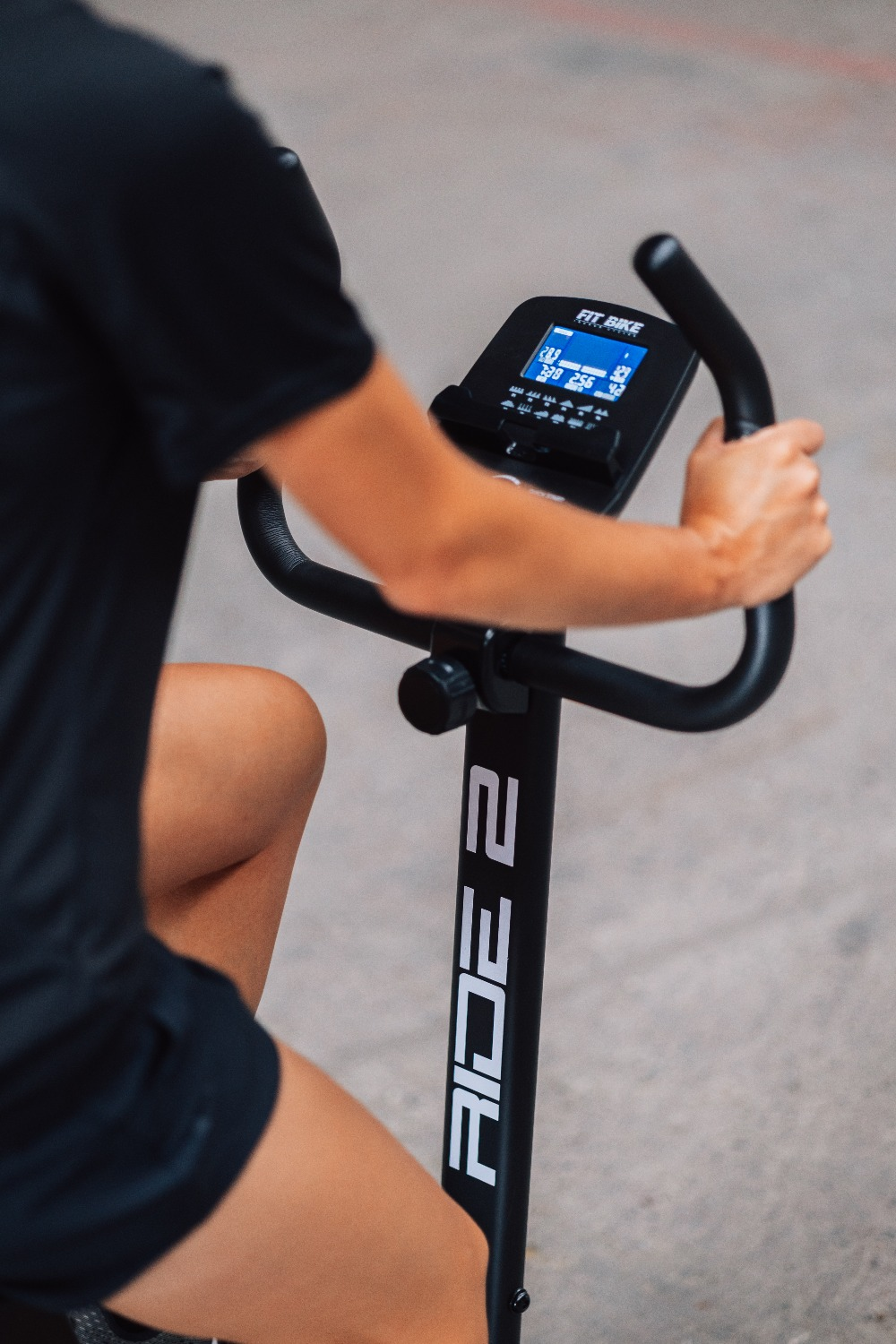 Person on a stationary bike, hands on handlebars. Bike has a digital display. Black bike and clothing.