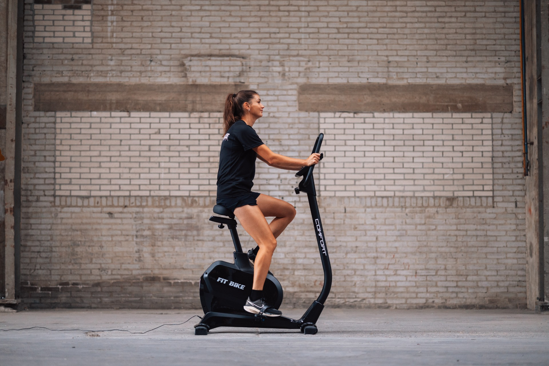 Vrouw op een hometrainer. Ze draagt ​​zwarte kleding en traint voor een bakstenen muur.
