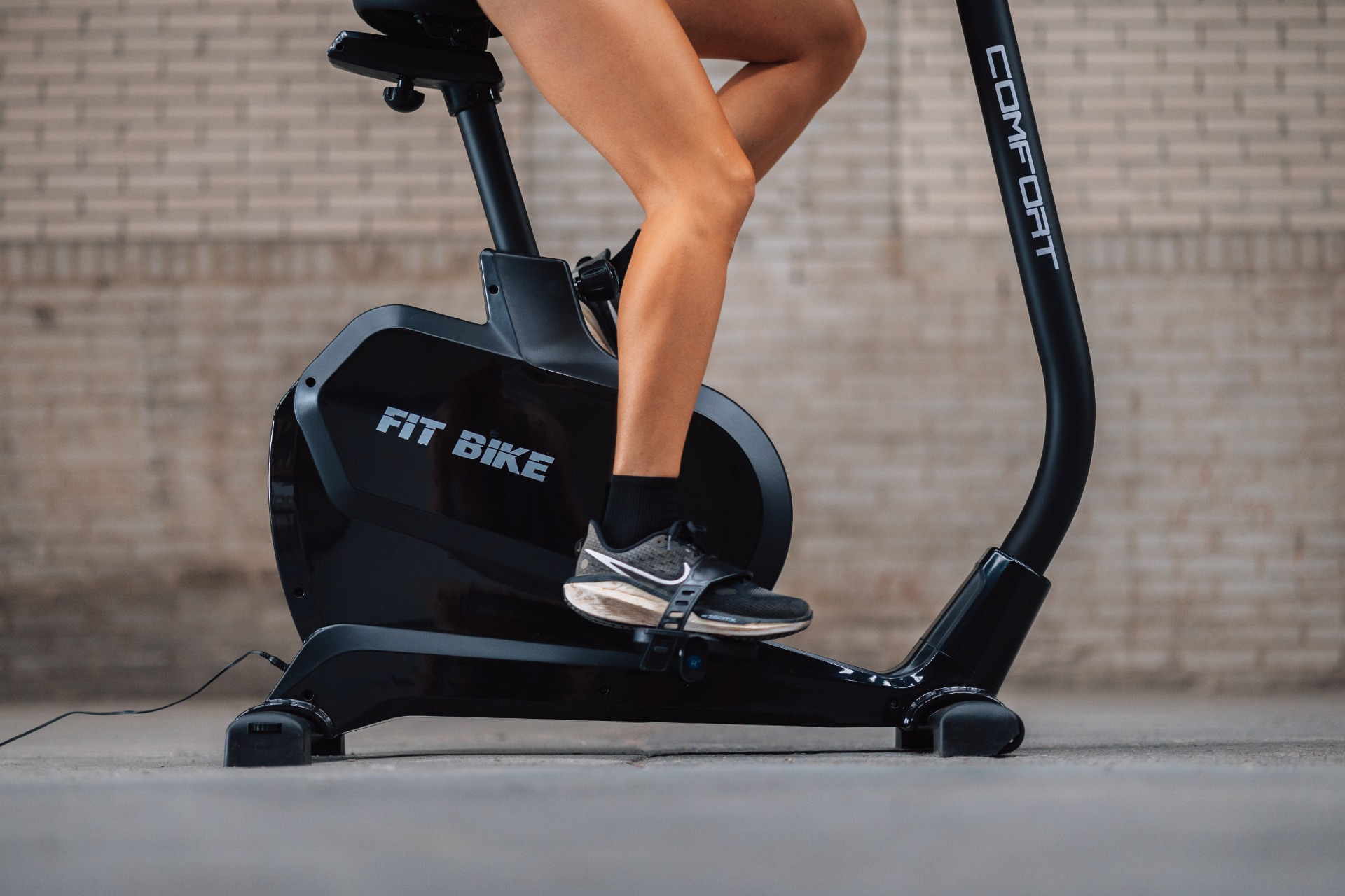 Een persoon fietst op een zwarte hometrainer. Op de fiets staat 'Fit Bike' en 'Comfort'. De benen en voeten van de persoon zijn zichtbaar.