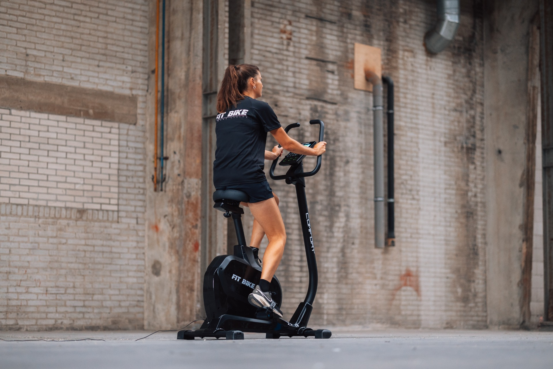 Vrouw op een zwarte hometrainer in een industriële omgeving. Ze draagt zwarte sportkleding. Achtergrond: bakstenen muur.