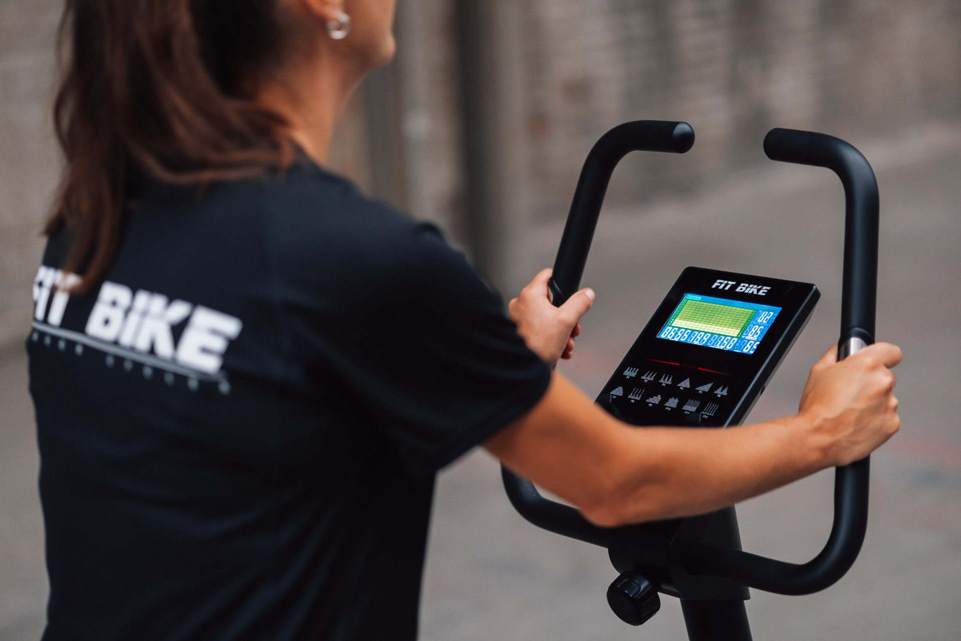 Een persoon gebruikt een Fit Bike. Het scherm van de fiets toont gegevens. De persoon draagt een zwart shirt met het Fit Bike logo.