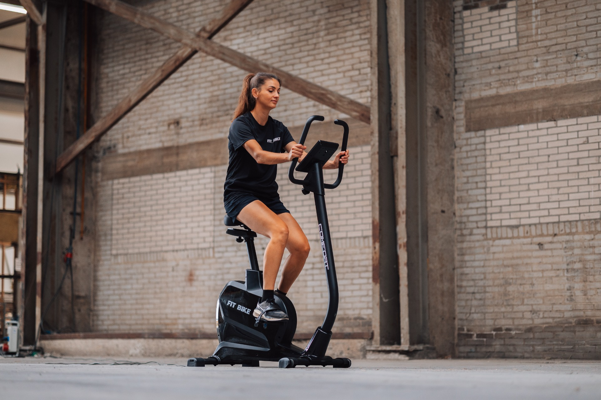 Een vrouw in het zwart traint op een hometrainer binnenshuis. De achtergrond toont bakstenen muren en houten balken.
