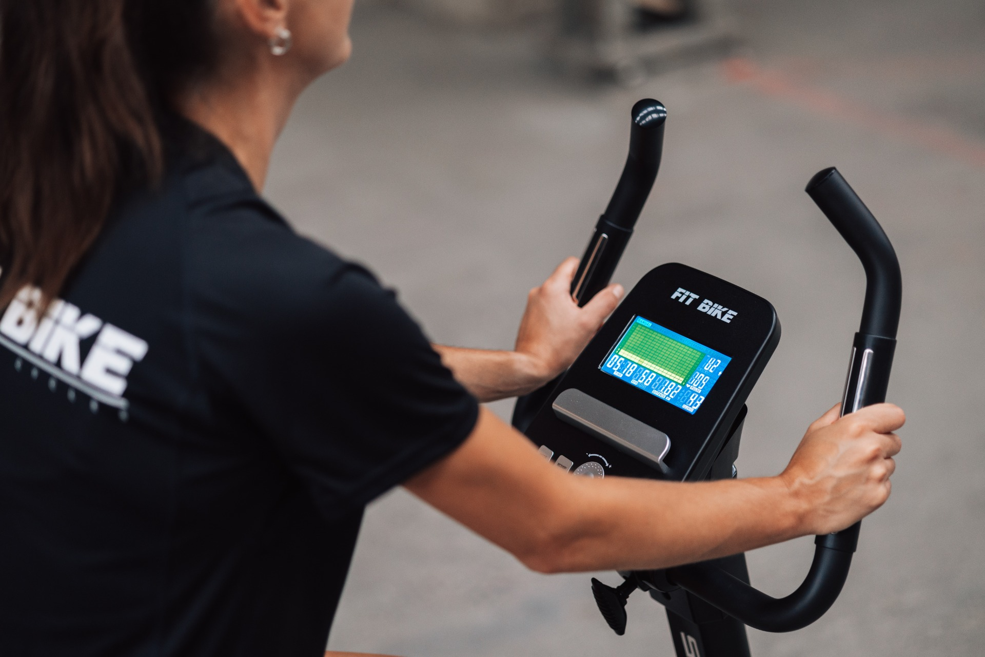 Femme utilisant un Fit Bike. Elle tient les poignées, l'écran affiche des statistiques. Le fond est flou.