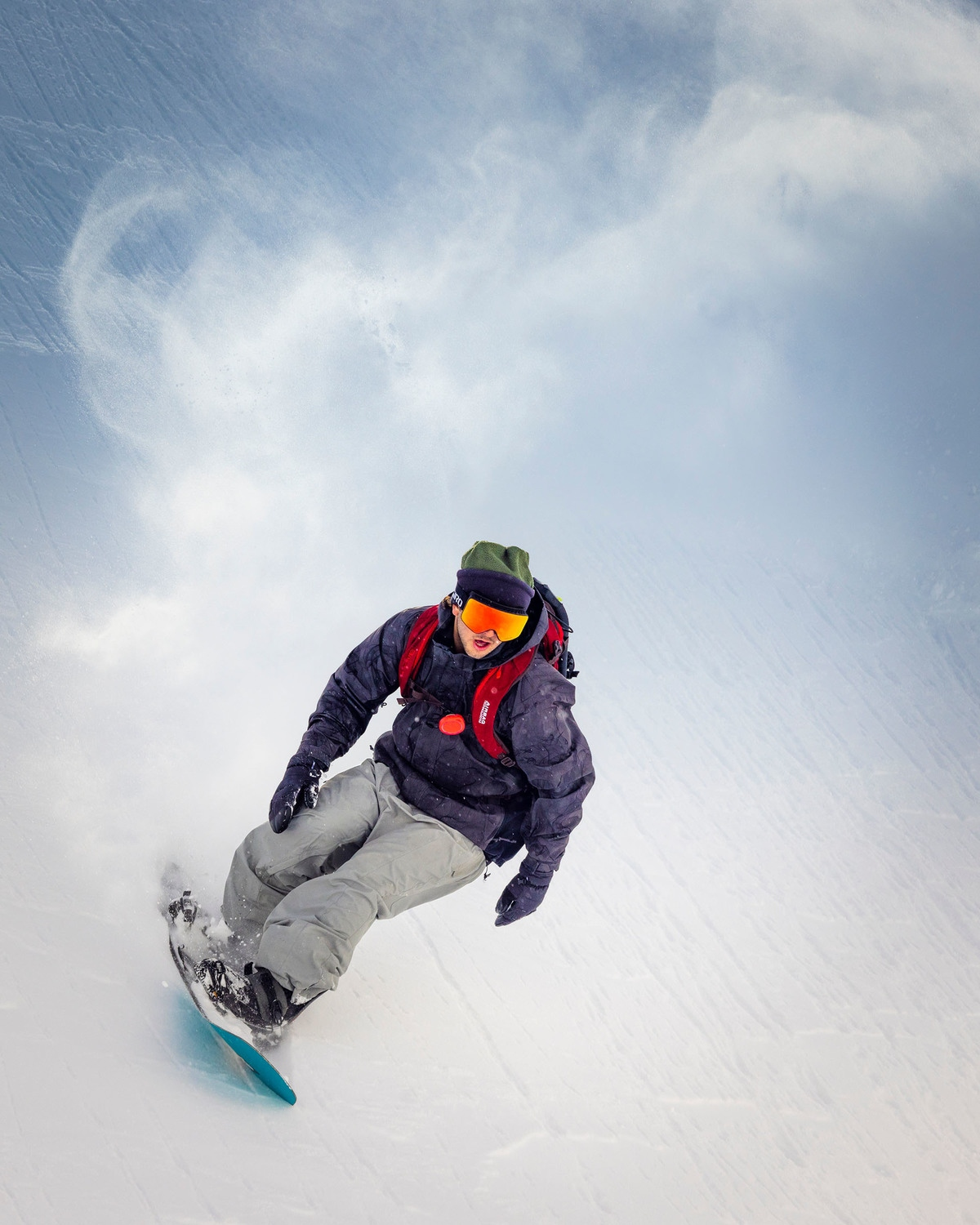 Un snowboarder desciende por una ladera nevada. Lleva chaqueta, pantalones grises y gafas naranjas. El cielo está nublado.