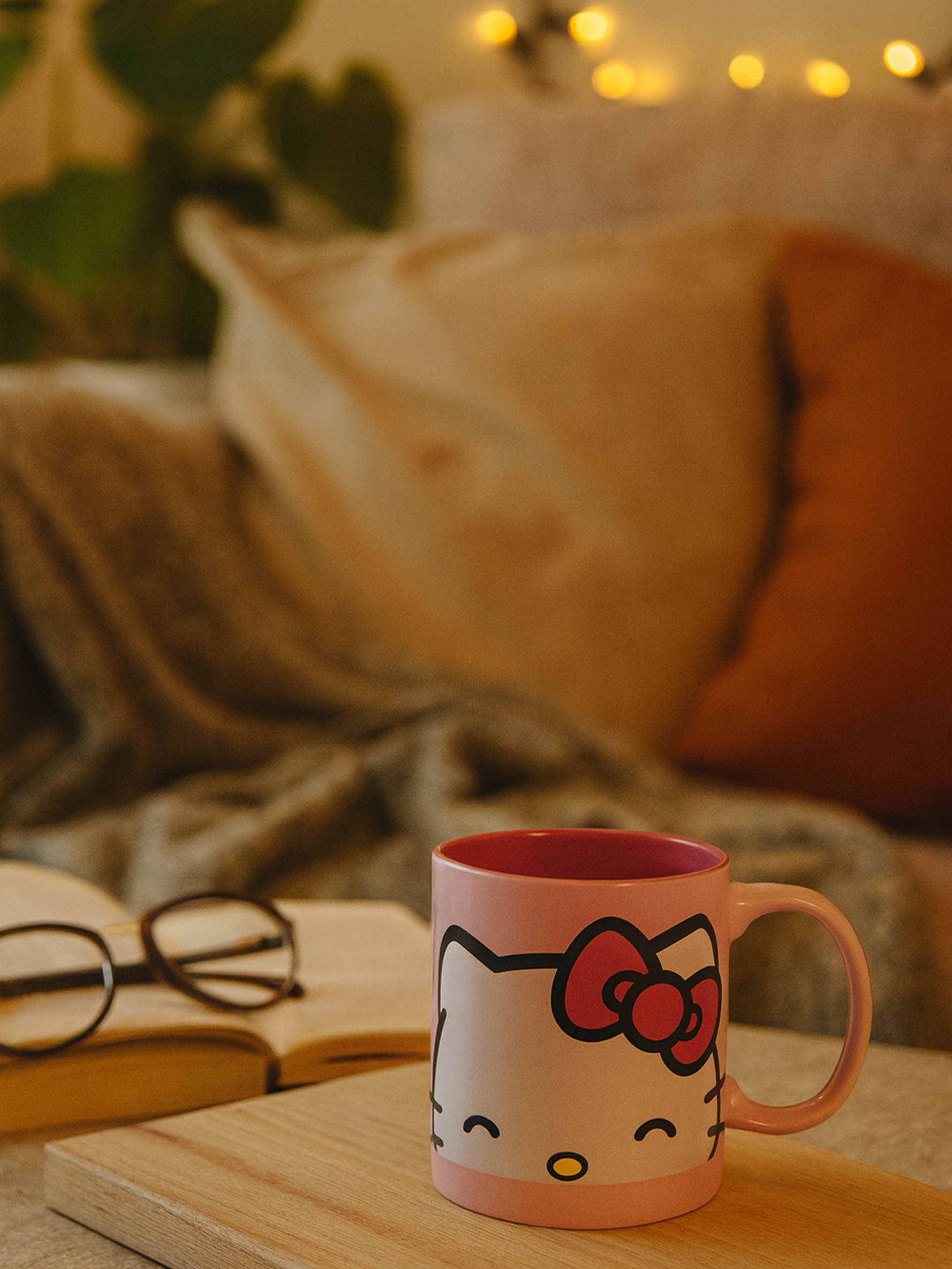 Eine rosa Hello Kitty-Tasse steht auf einem Holzbrett neben einem offenen Buch und einer Brille. Kissen und Lichter sind im Hintergrund.