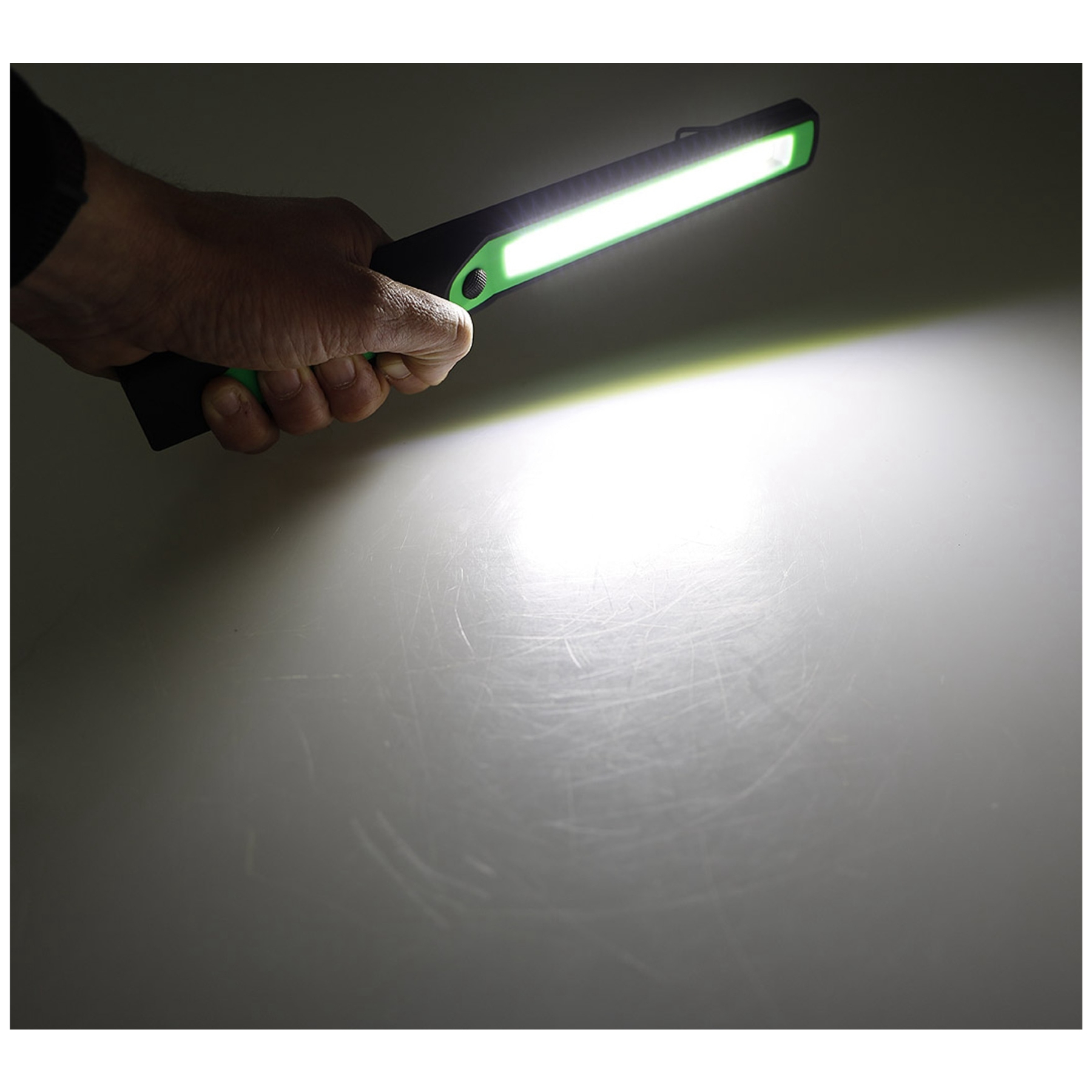 Eine Hand hält eine schwarz-grüne LED-Leuchte, die eine helle, weiße Oberfläche darunter beleuchtet. Das Licht ist an.