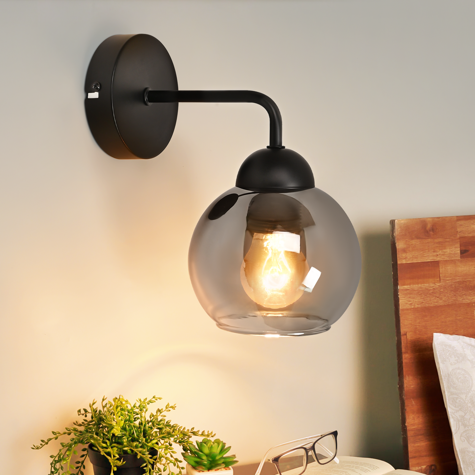 Applique nero con globo in vetro fumé. Una lampadina accesa all'interno. Un letto sullo sfondo. Piante e occhiali su un tavolo.